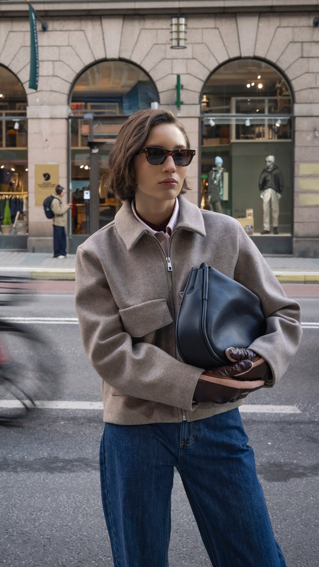 A woman wearing a beige wool zip-up jacket and sunglasses holds a black leather handbag on a city street.