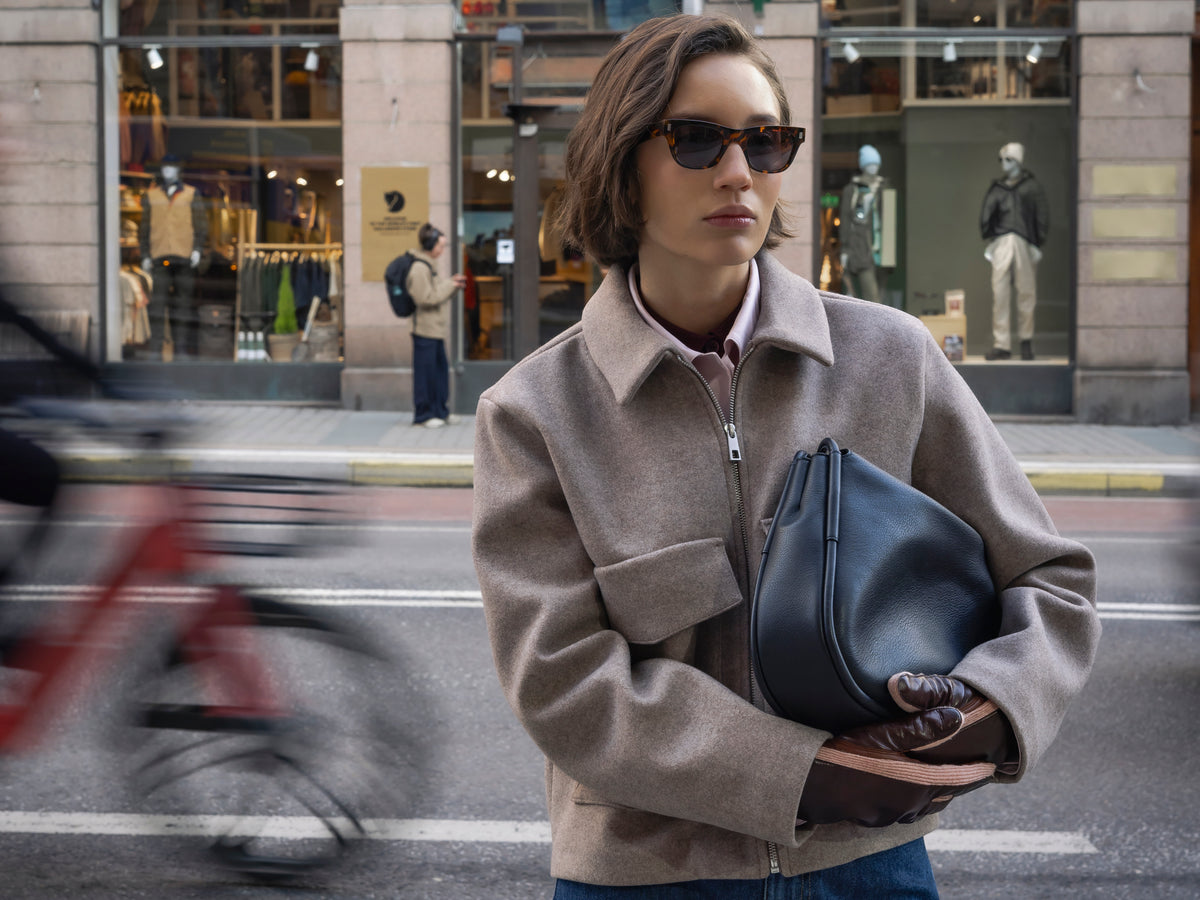 A woman wearing a beige wool zip-up jacket and sunglasses holds a black leather handbag on a city street.